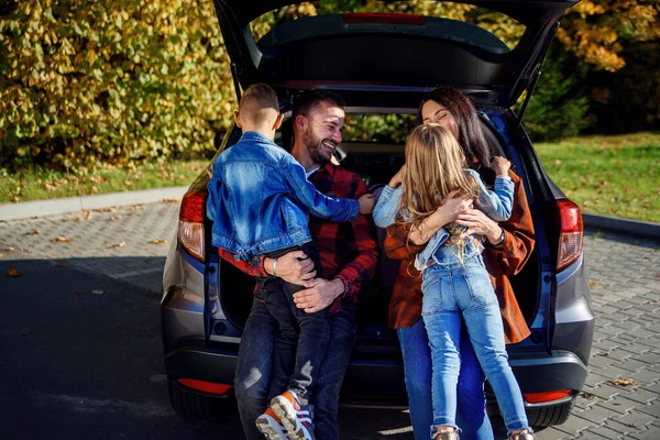 Felice allegro giovani genitori seduti nel bagagliaio auto e abbracciare i loro figli felici in esecuzione a loro . — Foto Stock