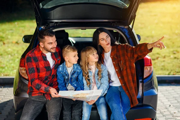 Familia satisfecha de padre guapo, madre bonita y niños encantadores que están mirando en la hoja de ruta mientras está sentado en el maletero de los coches en vacaciones familiares conjuntas. — Foto de Stock