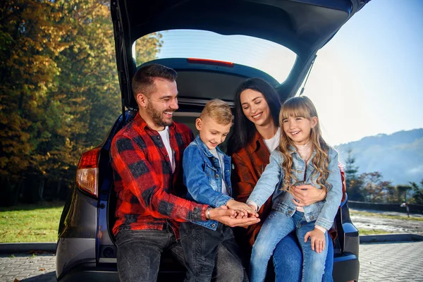 Genitori adorabili con i loro adorabili figli seduti nel bagagliaio della loro auto di famiglia, ridendo e dandosi cinque l'un l'altro. Godere felice concetto di tempo per la famiglia. — Foto Stock