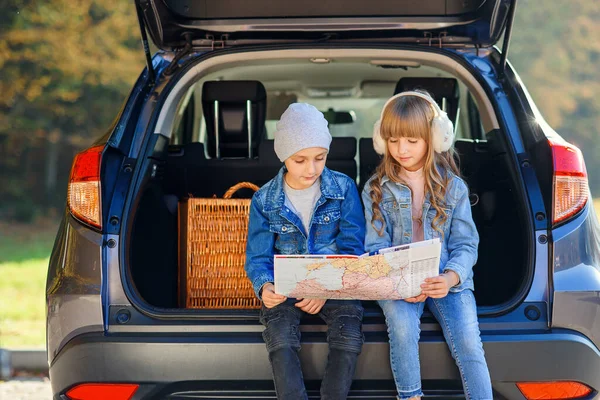 Милые мальчик и девочка смотрят на дорожную карту, сидя в багажнике автомобиля и обсуждая направление движения. Семейный отдых на автомобиле . — стоковое фото