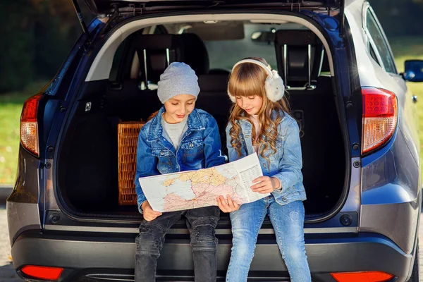 Agreeable boy and girl are looking at the road map while sitting in the autos trunk and discussing the move direction. Family vacation trip by car.