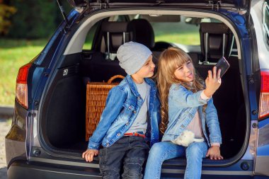 Şapkalı yakışıklı 10 yaşında bir çocuk ve telefonunda fotoğraf çeken, arabaların bagajında oturan uzun sarı saçlı güzel bir kız.