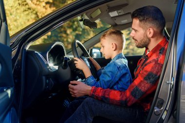 Yakışıklı baba, genç oğula araba sürmeyi öğretirken bir şeyi işaret ediyor.