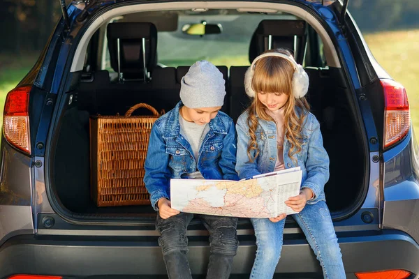 Agreeable boy and girl are looking at the road map while sitting in the autos trunk and discussing the move direction. Family vacation trip by car.