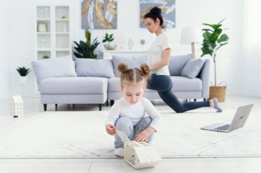 Sportif, sağlıklı annesi arka planda spor yaparken ve yoga egzersizleri yaparken evde oynayan komik bir bebek..
