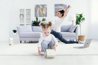 Sportif, sağlıklı annesi arka planda spor yaparken ve yoga egzersizleri yaparken evde oynayan komik bir bebek..