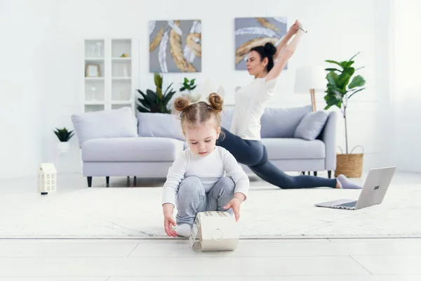 Grappig baby meisje spelen thuis, terwijl haar sportieve gezonde mama doet fitness en yoga oefeningen op de achtergrond. — Stockfoto