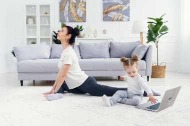 Komik kız laptopla oynarken sportif sağlıklı annesi evde yoga eğitimi alıyor..