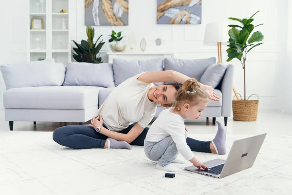 Αστείο κοριτσάκι παίζει με το laptop, ενώ σπορ υγιή μαμά της έχουν online γιόγκα κατάρτισης στο σπίτι. — Φωτογραφία Αρχείου