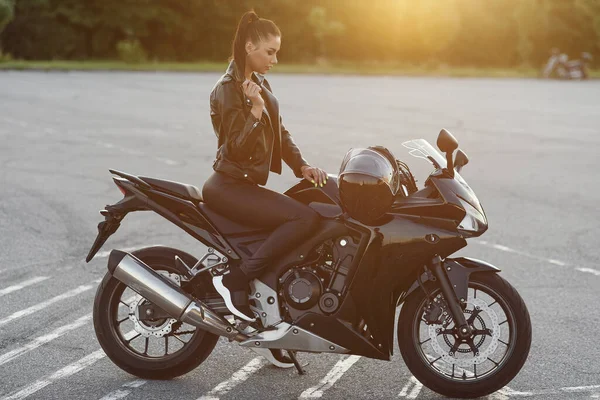 Attraktives Mädchen in schwarzer Lederjacke und Hose auf dem Outdoor-Parkplatz mit stylischem Sportmotorrad bei Sonnenuntergang. — Stockfoto