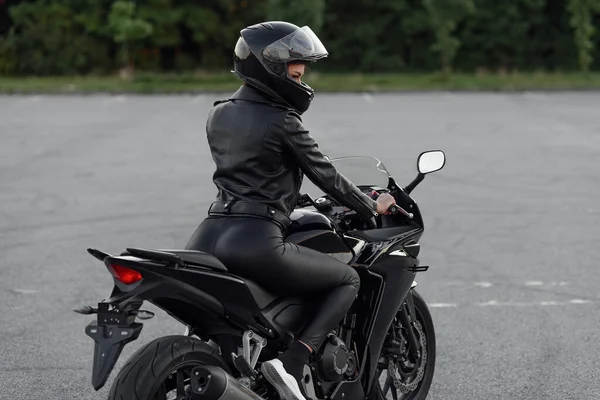 Jolie fille aux cheveux longs en veste et pantalon en cuir noir sur un parking extérieur avec une moto sport élégante au coucher du soleil. — Photo