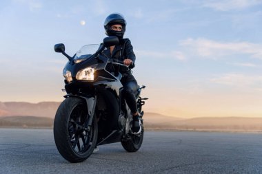 Uzun saçlı, siyah deri ceketli ve pantolonlu çekici bir kız gün batımında şık bir spor motosikletiyle açık havada park ediyor..