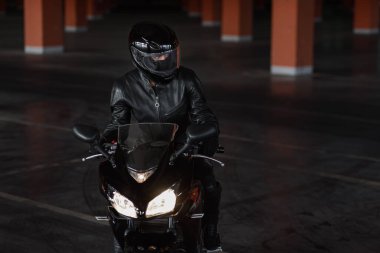 Siyah koruyucu üniformalı, eldivenli ve kasklı bir kız yeraltı otoparkında motosikletine biniyor..