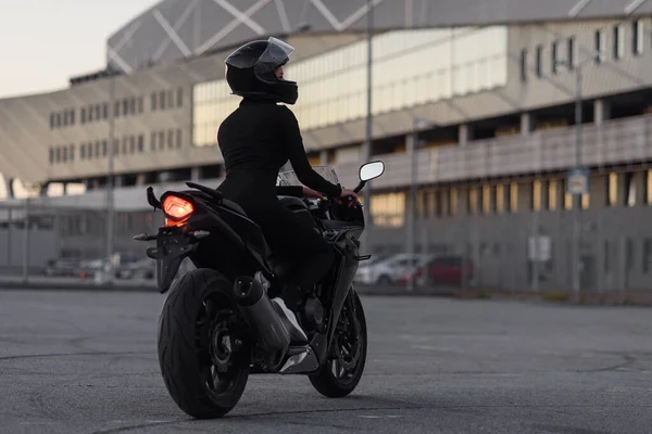 Tampilan belakang wanita muda yang menarik dalam setelan ketat hitam dan helm pelindung penuh pada sepeda motor olahraga di parkiran luar kota pada malam hari. — Stok Foto