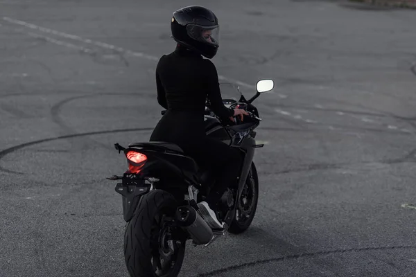 Attractive young woman in black tight-fitting suit and full-face protective helmet rides on sports motorcycle at urban outdoors parking in the evening.