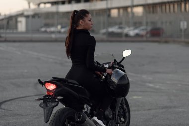 Siyah dar takım elbiseli çekici genç bir kadın ve tam yüz koruyucu kask takıyor. Spor motosikletleri, açık hava parkında akşam vakti..