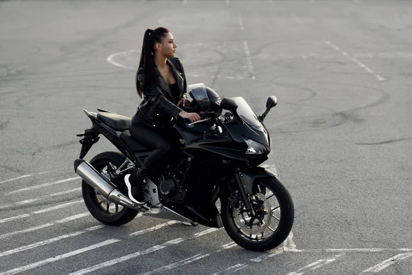 Gadis yang menarik dalam jaket kulit hitam dan celana di luar ruangan parkir dengan motor sport bergaya saat matahari terbenam. — Stok Foto