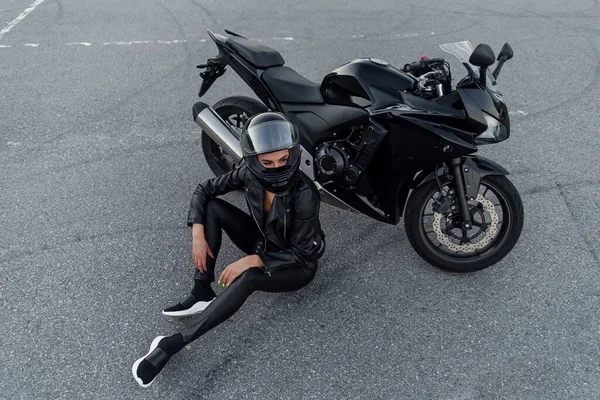 Mulher motociclista em jaqueta de couro preto e capacete de rosto cheio fica perto de motocicleta esportiva elegante no estacionamento urbano. Conceito de viagem e estilo de vida ativo . — Fotografia de Stock