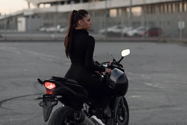 Attractive young woman in black tight-fitting suit and full-face protective helmet rides on sports motorcycle at urban outdoors parking in the evening. — Stock Photo, Image