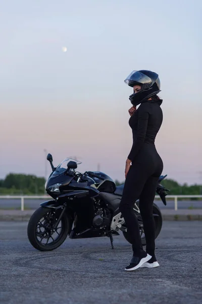 Wanita pengendara motor dengan jaket kulit hitam dan helm wajah penuh duduk di dekat motor sport bergaya di parkiran kota. Traveling dan konsep gaya hidup aktif. — Stok Foto