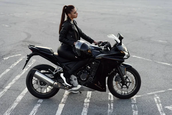 Menina atraente com cabelos longos em jaqueta de couro preto e calças no estacionamento ao ar livre com elegante moto esportiva ao pôr do sol. — Fotografia de Stock