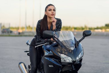 Uzun saçlı, siyah deri ceketli ve pantolonlu çekici bir kız gün batımında şık bir spor motosikletiyle açık havada park ediyor..