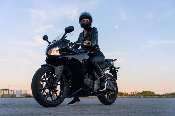 Attraente ragazza con i capelli lunghi in giacca di pelle nera e pantaloni sul parcheggio all'aperto con elegante moto sportiva al tramonto. — Foto Stock