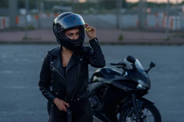 Mujer motorista en chaqueta de cuero negro y casco de cara completa se encuentra cerca de la motocicleta deportiva con estilo. Aparcamiento urbano, puesta de sol en la gran ciudad. Viajar y estilo de vida hipster activo. Las niñas poder . —  Fotos de Stock