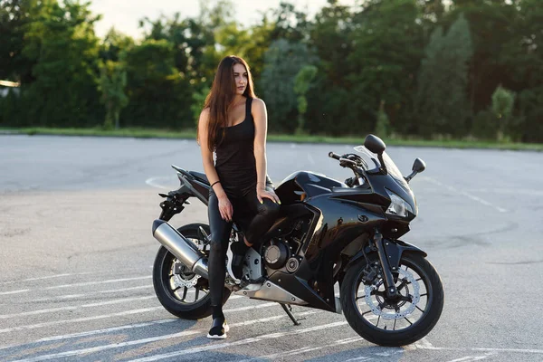 Attraktives Mädchen mit langen Haaren in schwarzer Lederjacke und Hose auf dem Außenparkplatz mit stylischem Sportmotorrad bei Sonnenuntergang. — Stockfoto