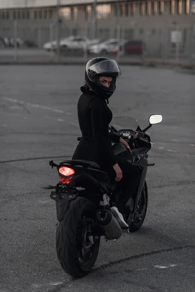 Rückenansicht einer attraktiven jungen Frau in schwarzem, eng anliegendem Anzug und Integralschutzhelm, die abends auf dem städtischen Außenparkplatz mit dem Sportmotorrad unterwegs ist. — Stockfoto