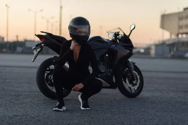 Mujer motorista en chaqueta de cuero negro y casco completo se sienta cerca de la motocicleta deportiva con estilo en el aparcamiento urbano. Concepto de viaje y estilo de vida activo . —  Fotos de Stock