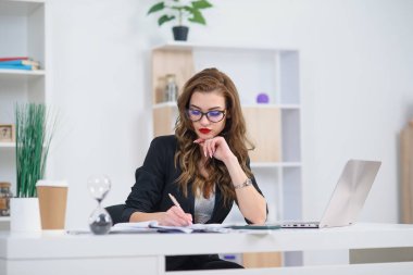 Ofisteki güzel iş kadını şık kıyafetler giyiyor, modern rahat ofisinde otururken kağıda bazı notlar yazıyor..