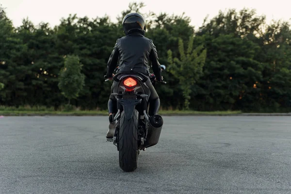 Attraktives Mädchen mit langen Haaren in schwarzer Lederjacke und Hose auf dem Außenparkplatz mit stylischem Sportmotorrad bei Sonnenuntergang. — Stockfoto