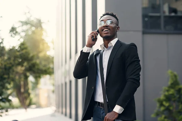 Joven empresario serio en traje negro de pie y hablando en el teléfono celular cerca del centro de negocios — Foto de Stock