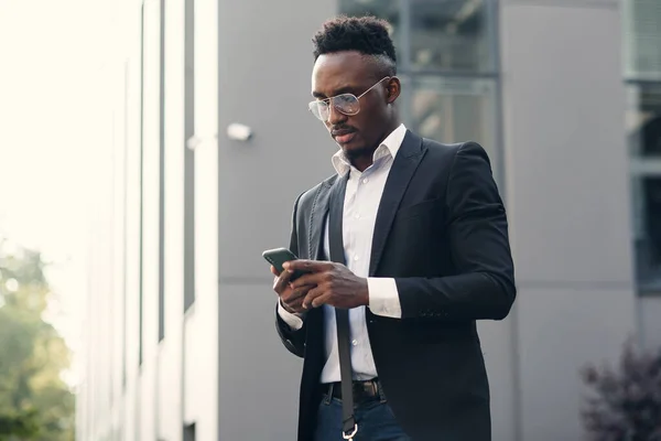 Serious negro empresario mensajes de texto en el teléfono inteligente al aire libre. Joven vendedor afroamericano que trabaja con móvil cerca del centro de negocios en el banco en el paisaje urbano — Foto de Stock