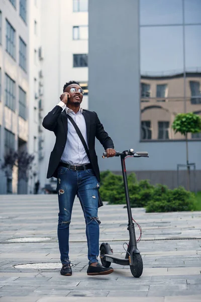 Hombre de negocios sonriente con scooter eléctrico parado frente al moderno edificio de negocios hablando por teléfono. — Foto de Stock