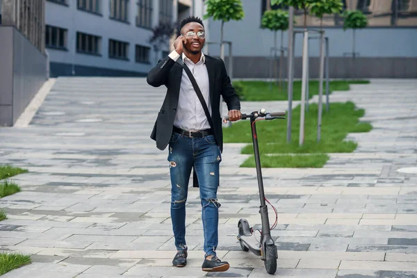 Hombre de negocios afroamericano sonriente con scooter eléctrico caminando cerca del moderno centro de negocios y hablando por teléfono. Tecnología moderna y concepto ecológico . — Foto de Stock