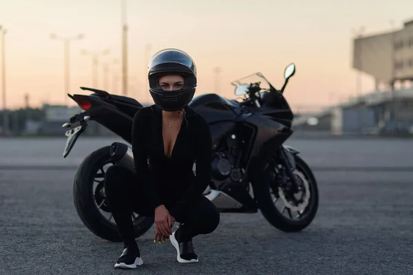 Mulher motociclista em jaqueta de couro preto e capacete de rosto cheio fica perto de motocicleta esportiva elegante no estacionamento urbano. Conceito de viagem e estilo de vida ativo . — Fotografia de Stock