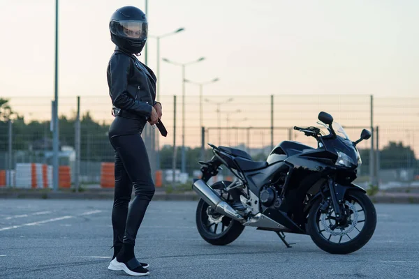 Mulher motociclista sexy em jaqueta de couro preto e capacete de rosto cheio fica perto de motocicleta esportiva elegante. Estacionamento urbano, pôr do sol na cidade grande. Viajando e estilo de vida hipster ativo . — Fotografia de Stock
