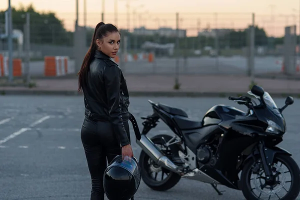 Back view of beautiful girl in black leather jacket and pants holding full face helmet on outdoors parking with stylish sports motorcycle at sunset. Freedom lifestyle concept. — Stock Photo, Image