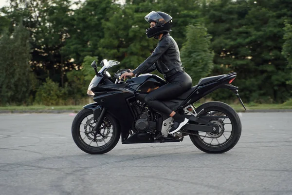 Elegante donna in giacca di pelle nera, pantaloni e casco protettivo integrale cavalca su moto sportive al parcheggio urbano all'aperto. — Foto Stock