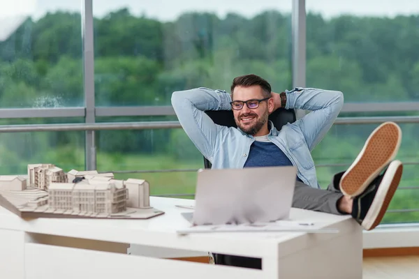 Happy office worker in casual clothes laid his feet on workspace table while dreaming about rest or vacations. Joyful architect relax on the workplace successfully completing his project.