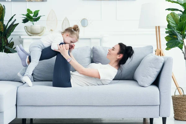 Feliz alegre jovem mãe e sua filha criança rindo juntos e se divertindo deitado no sofá. Mãe alegre abraço brincando com sua menina bonita em casa criança se divertindo relaxando com a mãe no sofá . — Fotografia de Stock