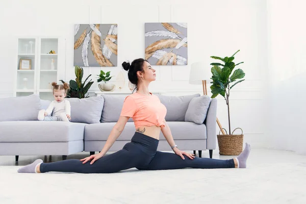 Mãe calma feliz fazendo exercícios matinais em pose de ioga enquanto sua filhinha brincando em casa . — Fotografia de Stock