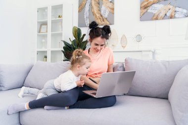 Genç, güzel anne ve küçük mutlu kızı evdeki kanepede otururken dizüstü bilgisayar kullanıyorlar ve gülümsüyorlar.