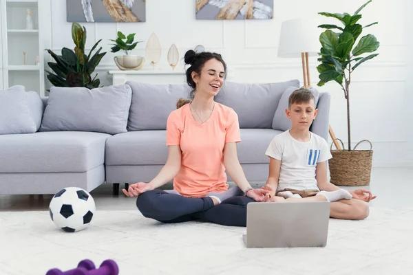 Mãe e filho praticam ioga com laptop no chão. Mãe esportiva com crianças se exercitando de manhã em casa. — Fotografia de Stock