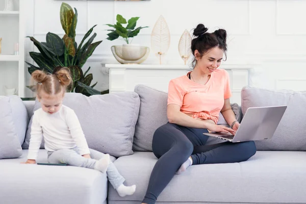 Jovem mãe bonita e sua pequena filha feliz estão usando um laptop e sorrindo enquanto estão sentados no sofá em casa — Fotografia de Stock