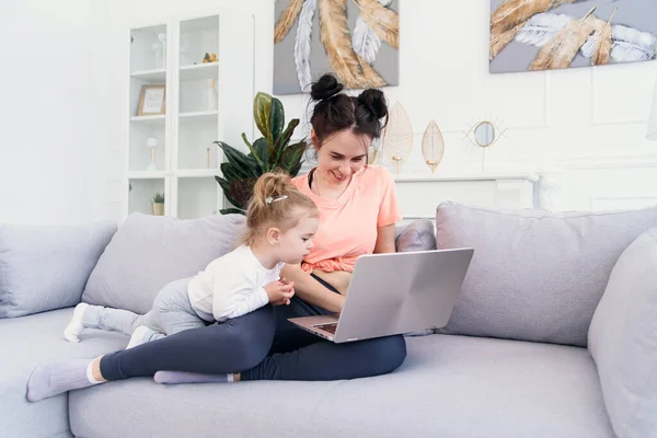 Jovem mãe bonita e sua pequena filha feliz estão usando um laptop e sorrindo enquanto estão sentados no sofá em casa — Fotografia de Stock