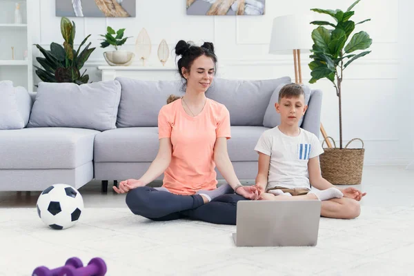 Mãe e filho praticam ioga com laptop no chão. Mãe esportiva com crianças se exercitando de manhã em casa. — Fotografia de Stock