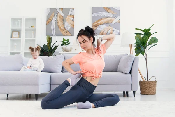 Bela mãe faz exercícios de alongamento matinal enquanto sua filhinha brincava em casa. Conceito de estilo de vida saudável. — Fotografia de Stock